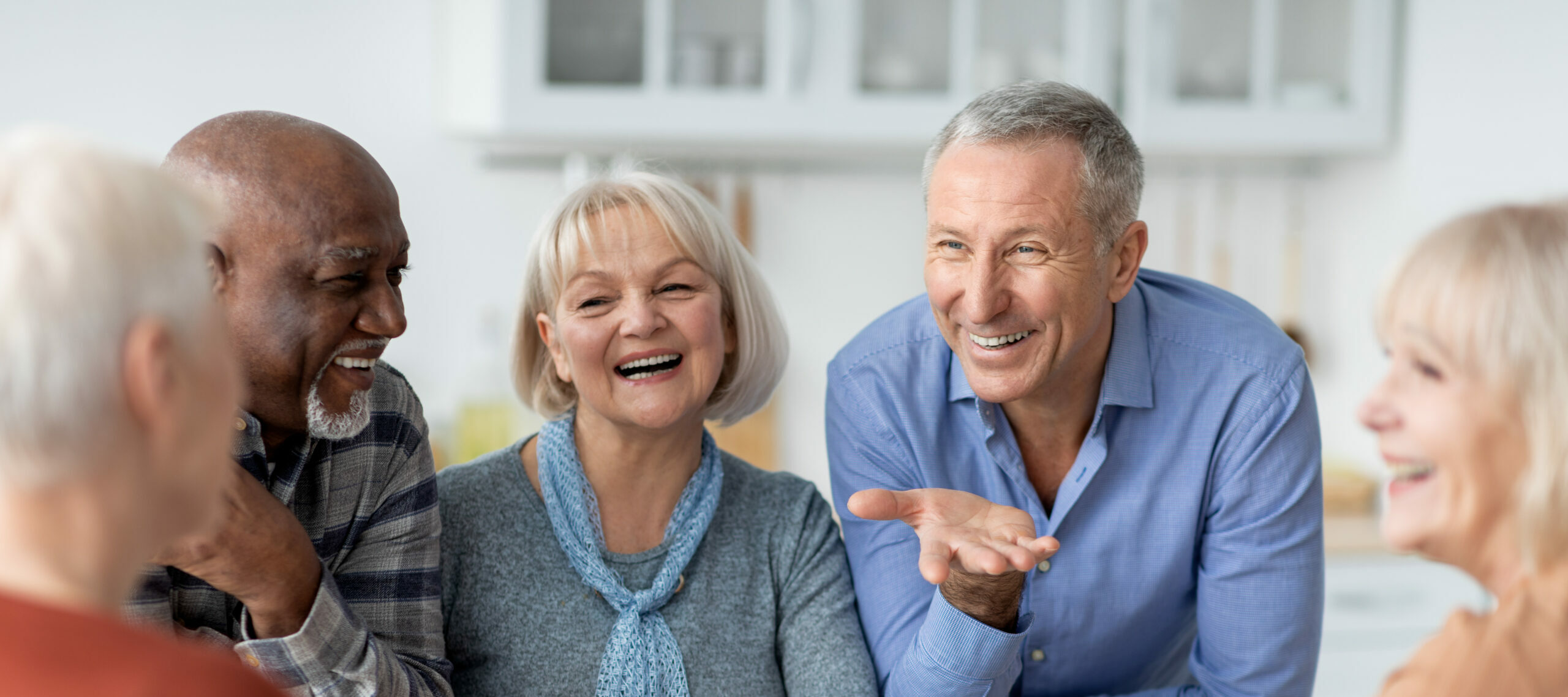 Multiracial group of happy senior people sitting around table drinking tea with cake and having conversation, smiling and laughing, having home party or enjoying time at nursing home